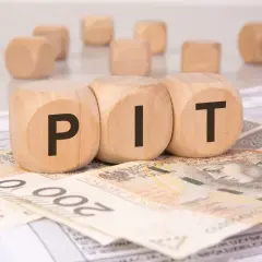 Drewniane kostki z literami "PIT" leżą na banknotach i dokumentach. Może to symbolizować rozliczenie podatkowe lub umowę darowizny.