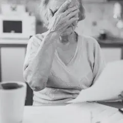 Starsza kobieta z kubkiem kawy w dłoni, zasłaniająca twarz, analizuje dokumenty. Zrid.