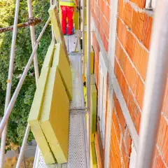 Prace budowlane: montaż izolacji na rusztowaniu. Trwa modernizacja budynku, widać ceglaną ścianę i żółte płyty izolacyjne.