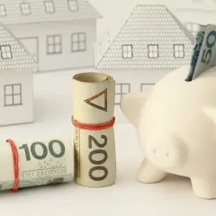Zabezpieczenie domu: czy zadatek jest zwrotny? Zwinięte banknoty 100 i 200 zł, świnka-skarbonka z banknotem 50 zł i białe modele domów.