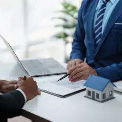 Dwóch mężczyzn w garniturach omawia dokumenty przy biurku z modelem domu i laptopem. Rozmowa dotyczy opłaty adiacenckiej.