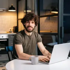 Uśmiechnięty mężczyzna z kręconymi włosami pracuje na laptopie w kuchni, planując, jak zapłacić czynsz.