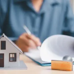 Model domu i pieczątka obok dokumentów, na których ktoś pisze. To symbol finalizacji umowy deweloperskiej.