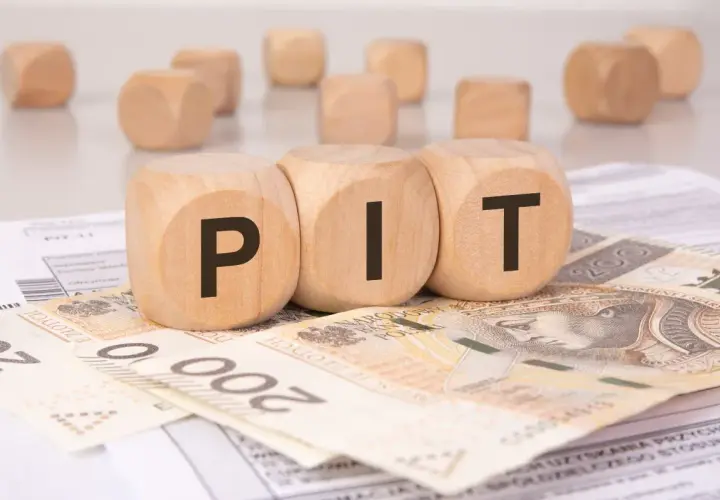 Drewniane kostki z literami "PIT" leżą na banknotach i dokumentach. Może to symbolizować rozliczenie podatkowe lub umowę darowizny.