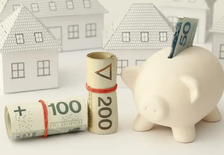 Zabezpieczenie domu: czy zadatek jest zwrotny? Zwinięte banknoty 100 i 200 zł, świnka-skarbonka z banknotem 50 zł i białe modele domów.