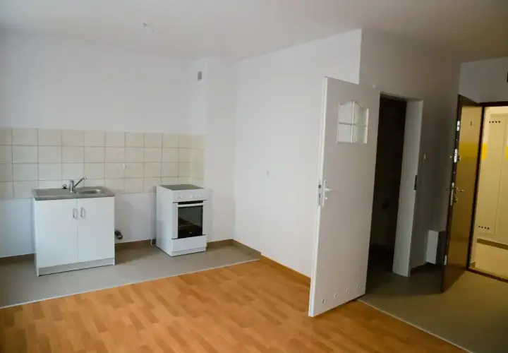 Proste mieszkanie socjalne z aneksem kuchennym: zlew, kuchenka i drzwi otwarte na korytarz.