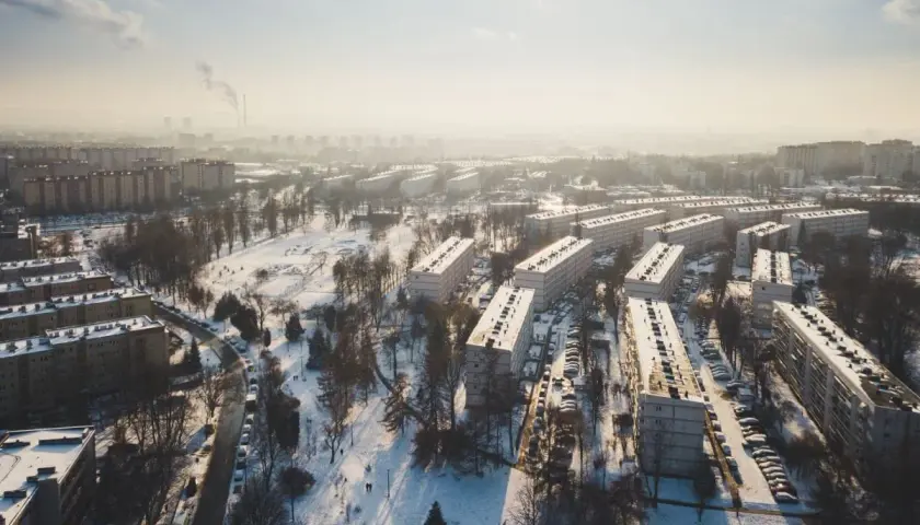 Zimowy krajobraz Krakowa z blokami mieszkalnymi i parkiem. Widać dymiące kominy w oddali, co może wpływać na ceny mieszkań w Krakowie.
