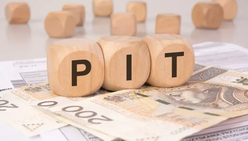 Drewniane kostki z literami "PIT" leżą na banknotach i dokumentach. Może to symbolizować rozliczenie podatkowe lub umowę darowizny.