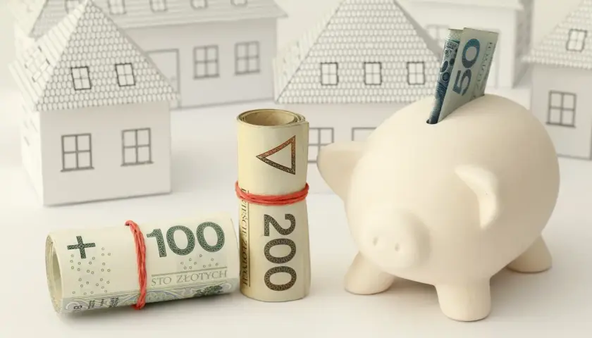 Zabezpieczenie domu: czy zadatek jest zwrotny? Zwinięte banknoty 100 i 200 zł, świnka-skarbonka z banknotem 50 zł i białe modele domów.