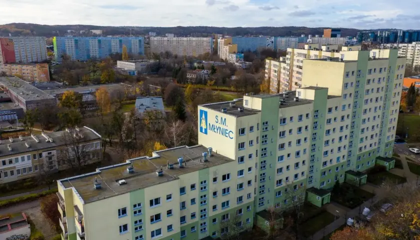 Duży blok mieszkalny S.M. Młyniec z charakterystycznym logo, otoczony innymi budynkami i drzewami jesienią.