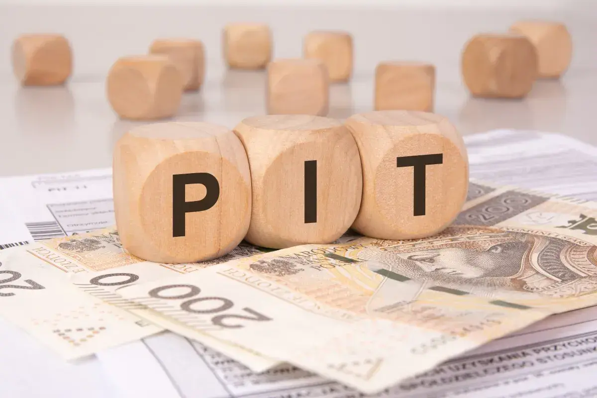 Drewniane kostki z literami "PIT" leżą na banknotach i dokumentach. Może to symbolizować rozliczenie podatkowe lub umowę darowizny.