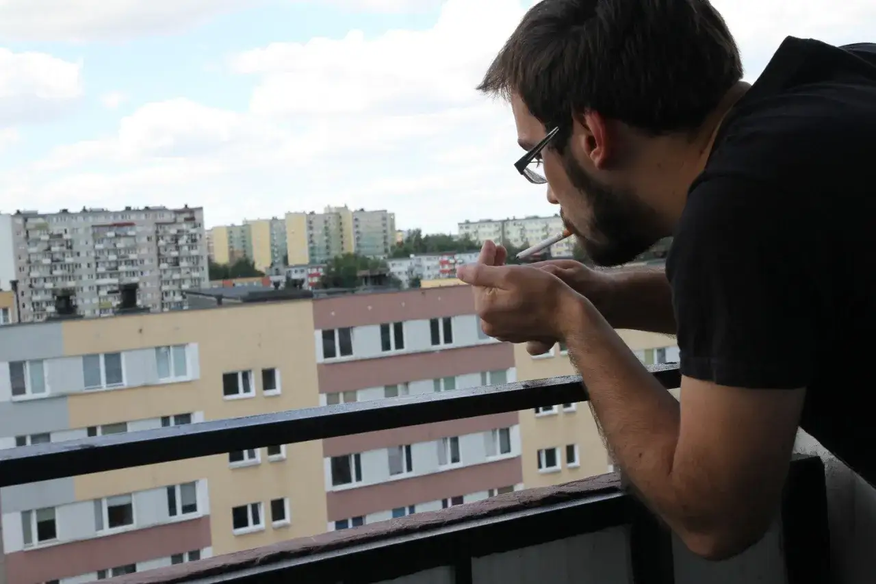 Mężczyzna z papierosem na balkonie, nad blokami. Czy można palić na balkonie? To pytanie zadaje sobie wielu mieszkańców.