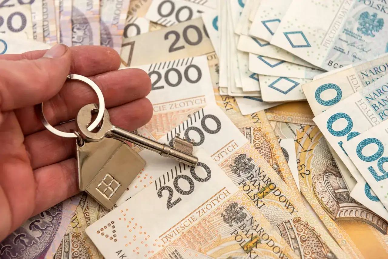 Dłoń trzyma klucz z breloczkiem w kształcie domu na tle polskich banknotów. Symbolizuje to zakup nieruchomości lub dziedziczenie, gdzie ważny jest zachowek.